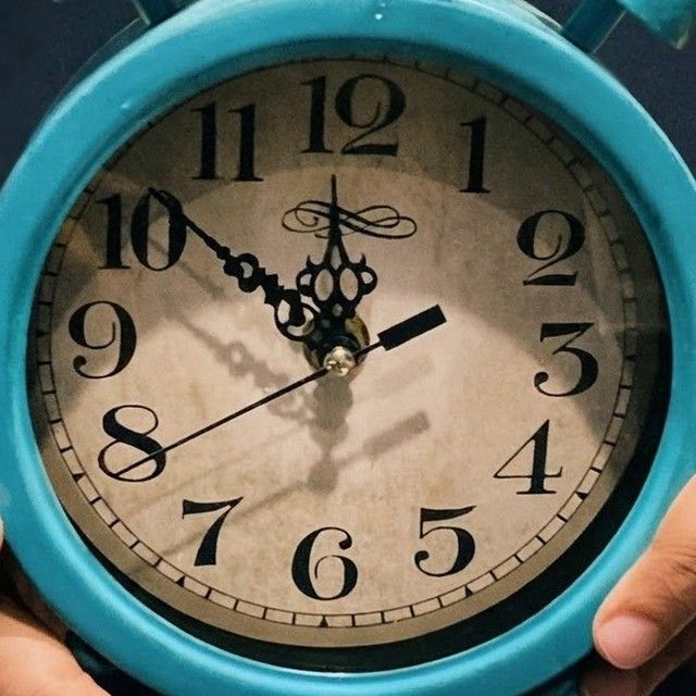 A hand holding a teal alarm clock close to the lens